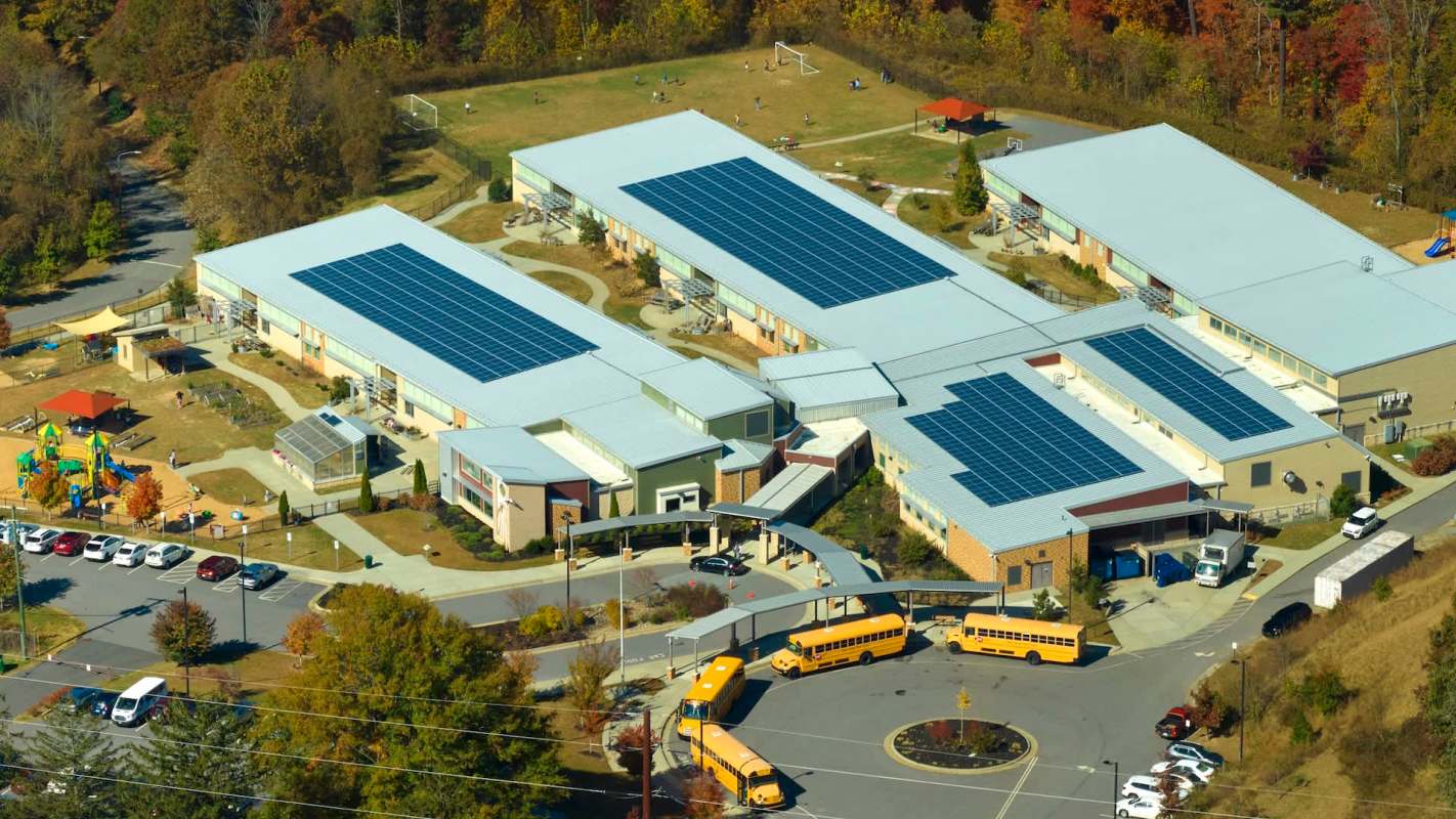 A school building with solar panels on the roof, surrounded by a playground and parked school buses.
