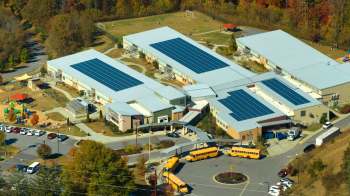 A school building with solar panels on the roof, surrounded by a playground and parked school buses.