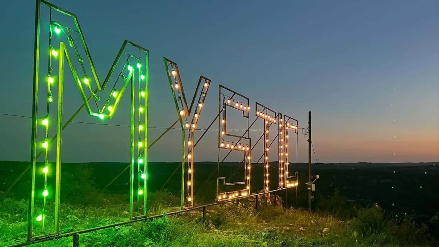 A neon sign spelling "MYSTIC" set against a twilight sky with a view of distant hills.