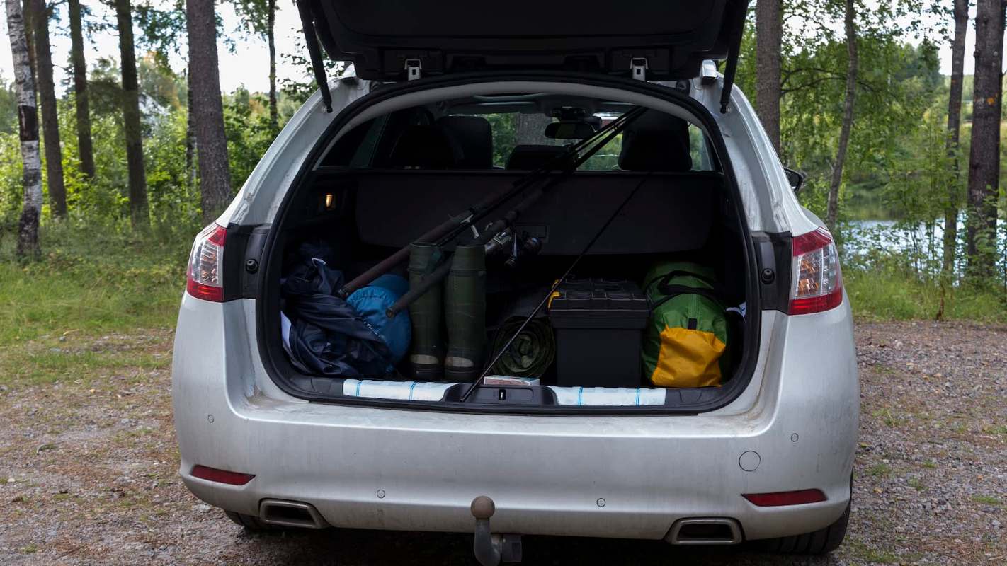 The trunk of a car is filled with fishing gear, boots, a green bag, and a yellow bag, surrounded by trees.