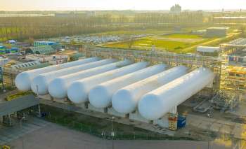Several large white storage tanks are positioned in an industrial area, with buildings and vehicles nearby.