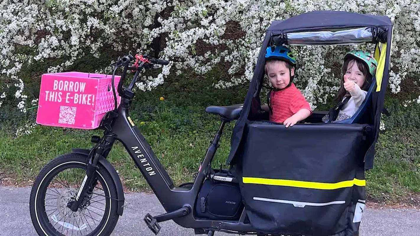 Two children sit in an e-bike trailer next to a bicycle with a pink box labeled "BORROW THIS E-BIKE!" and flowers in the background.