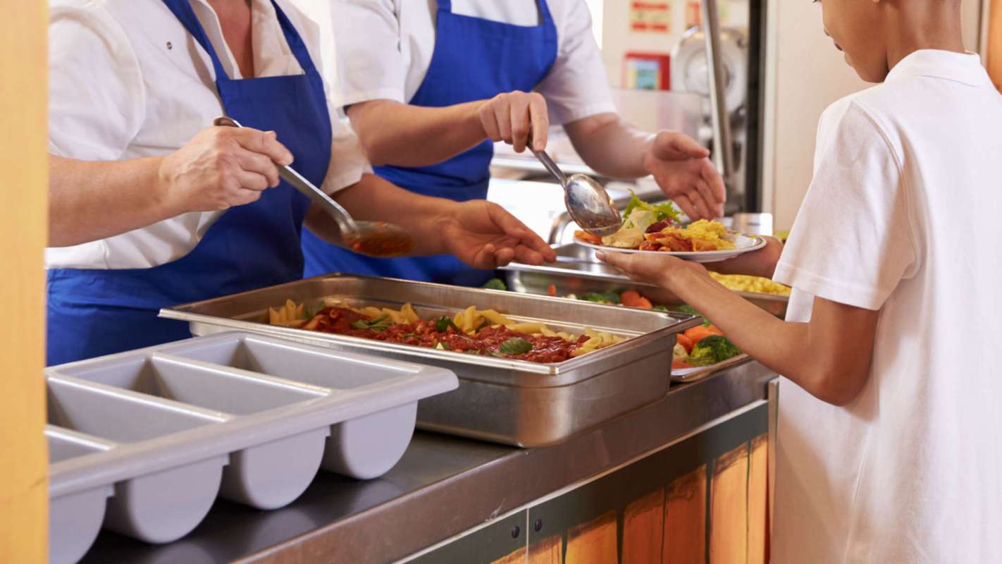 A child receives a plate of food from servers in a cafeteria.
