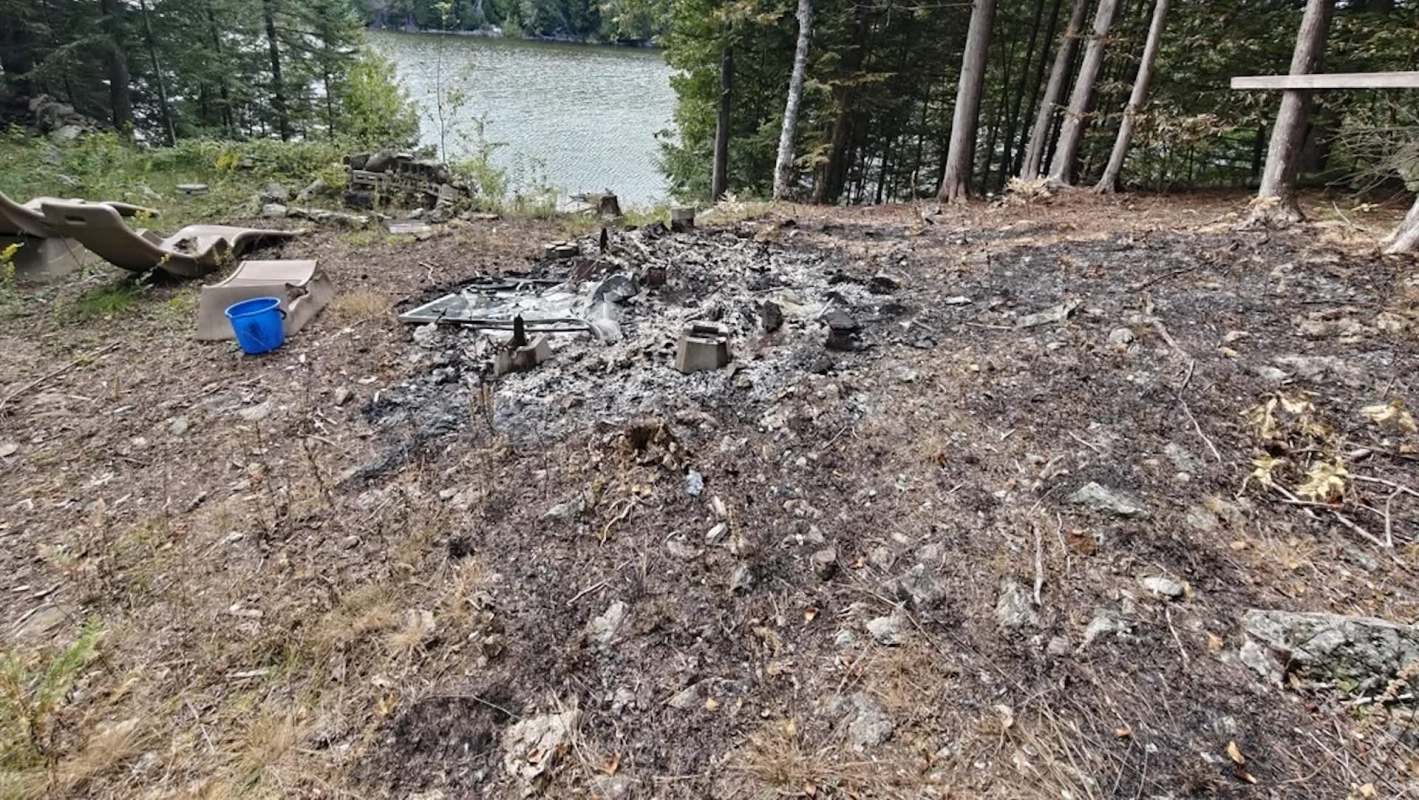 A burned area with remnants of a campfire and scattered debris near a body of water and forested surroundings.