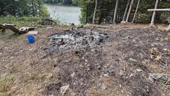 A burned area with remnants of a campfire and scattered debris near a body of water and forested surroundings.