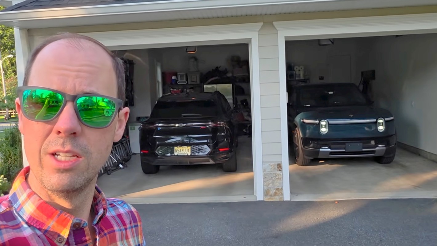A man stands outside a garage with two parked electric vehicles, one black Chevy SUV and one green Rivian.