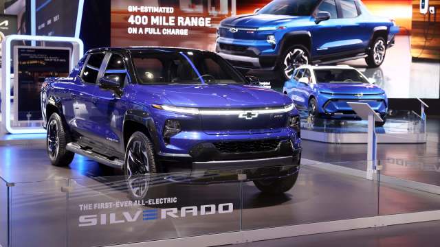 A blue Chevrolet Silverado electric truck displayed at an auto show, with another vehicle in the background.