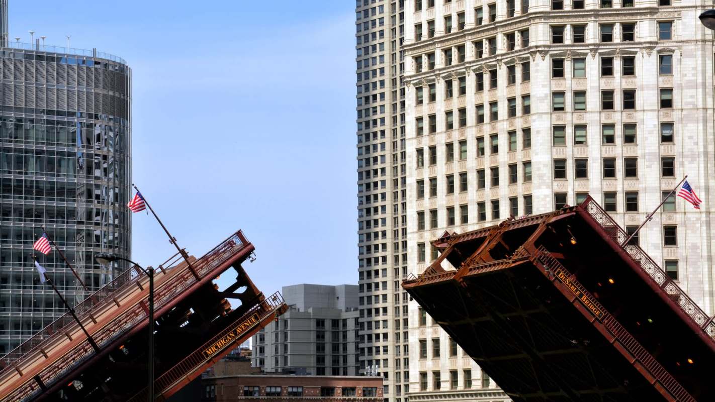 A drawbridge is raised, with tall buildings and American flags in the background.