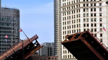 A drawbridge is raised, with tall buildings and American flags in the background.
