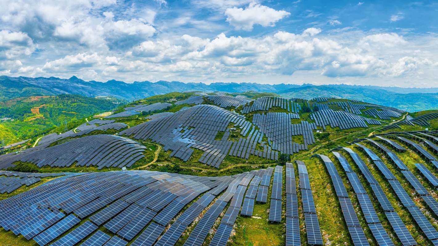 A vast solar panel installation on rolling hills with mountains in the background under a partly cloudy sky.