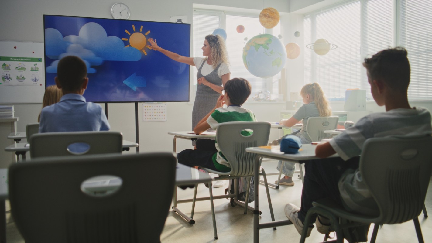 A teacher points to a screen displaying a weather illustration in a classroom filled with students.