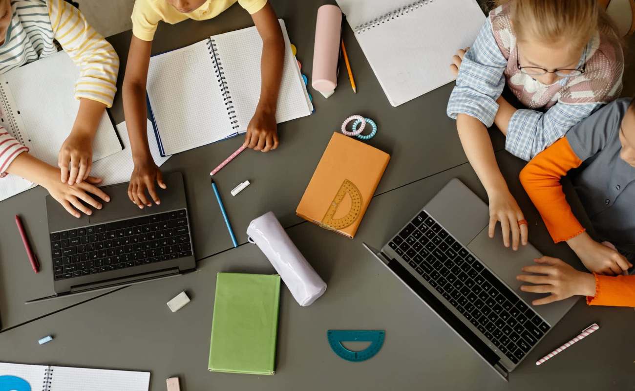 Several hands reach toward laptops and notebooks on a table, surrounded by school supplies.