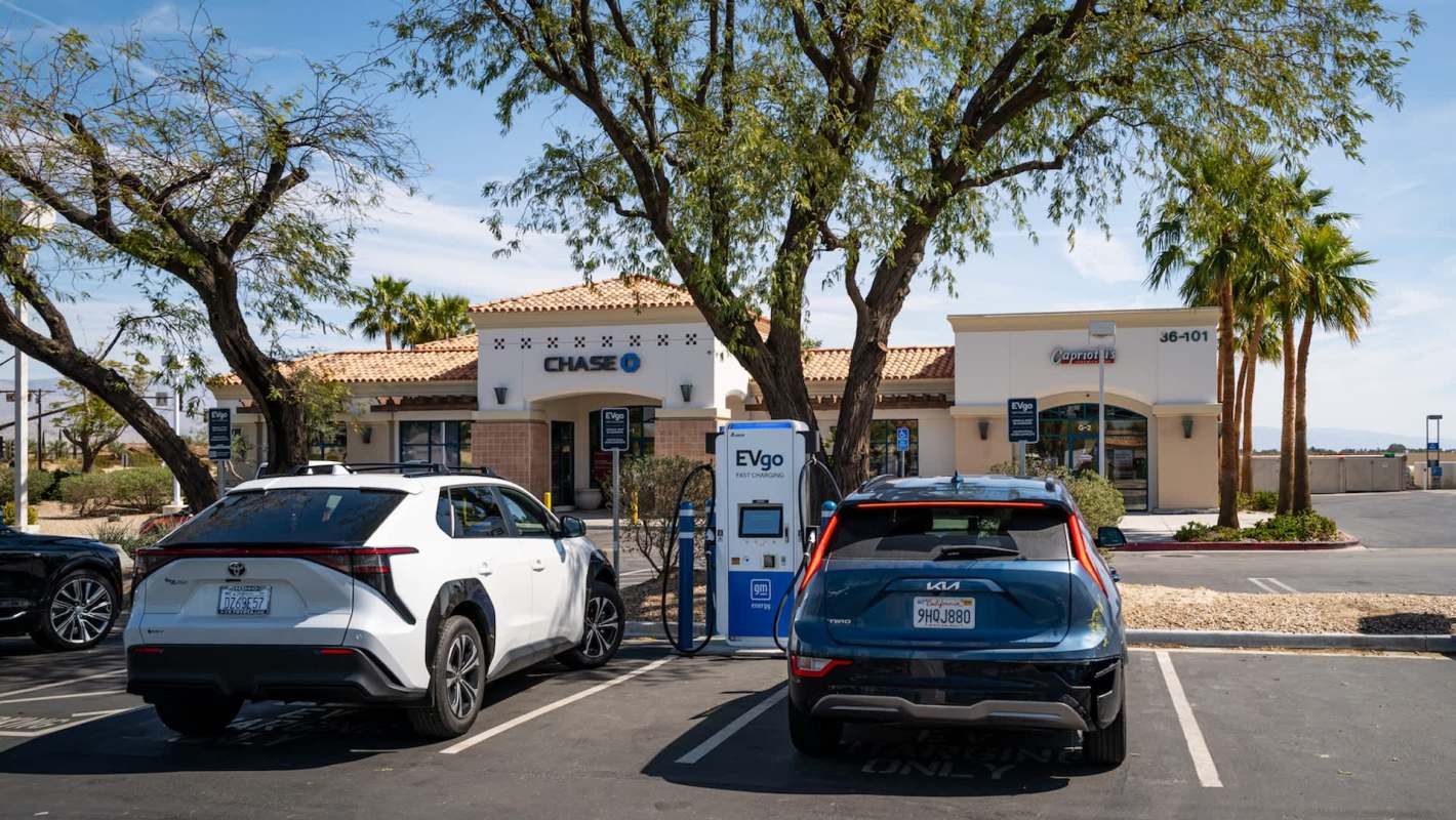 Two electric vehicles parked at a charging station outside a bank and a restaurant, surrounded by trees.
