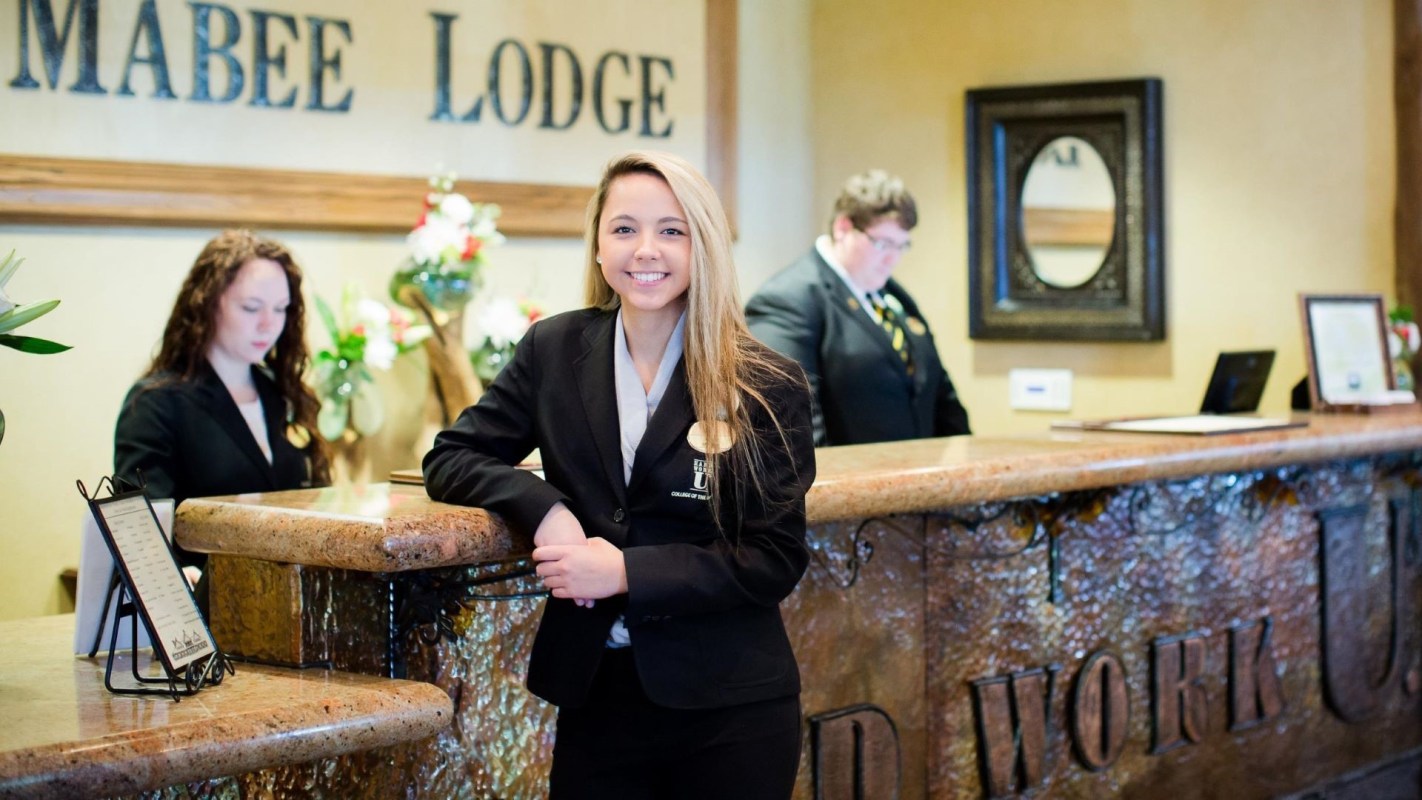 A receptionist stands at a hotel front desk with a coworker in the background.