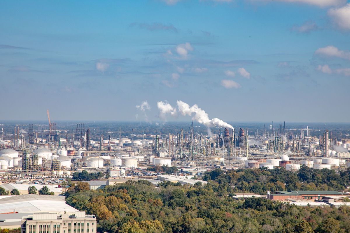 An industrial landscape featuring storage tanks, smoke stacks, and plumes of smoke against a blue sky.