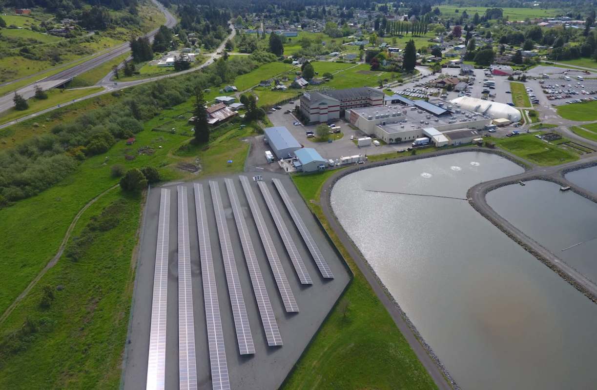An aerial view of solar panels near a building and a pond in a rural area with greenery and roads.