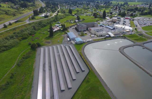 An aerial view of solar panels near a building and a pond in a rural area with greenery and roads.