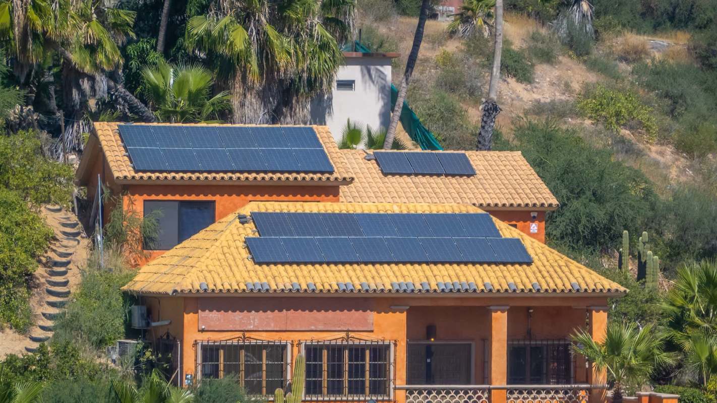 A house with a terracotta roof and solar panels, surrounded by palm trees and cacti.