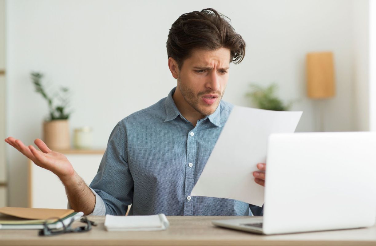 A man looks confused at a sheet of paper while seated at a desk with a laptop.