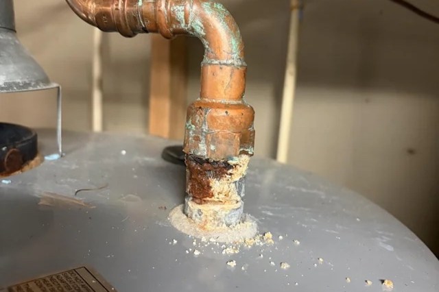 A corroded copper pipe connected to a water heater with rust and debris around the base.