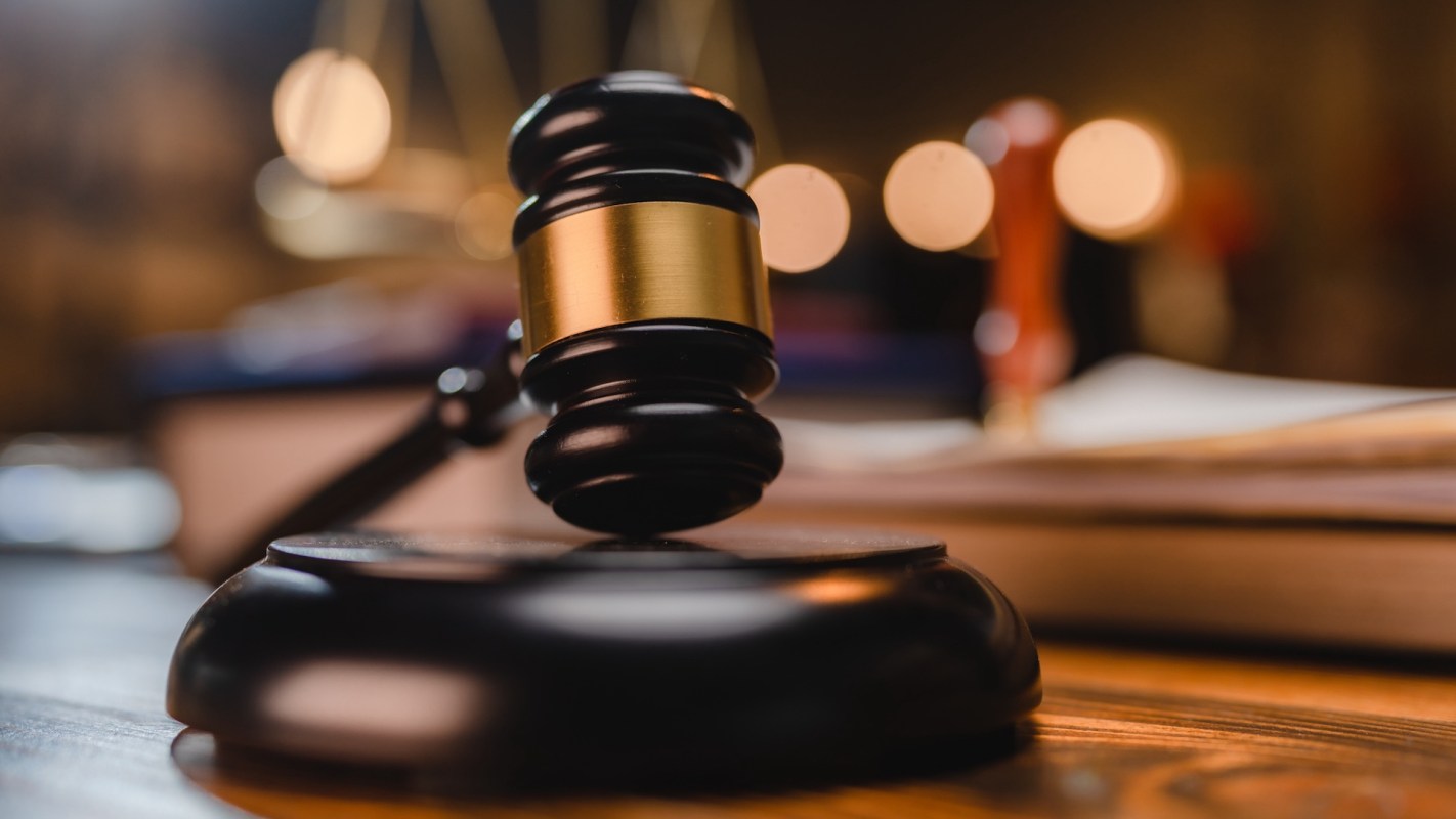 A gavel rests on a wooden surface, with a blurred background of legal books and scales.