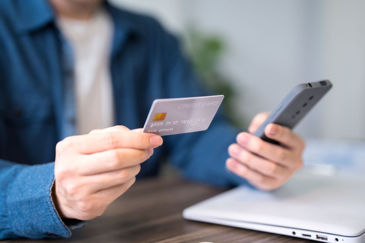 A person holds a credit card in one hand and a smartphone in the other, sitting at a table with a laptop.
