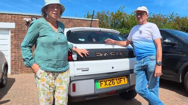 A woman and a man stand next to a white Dacia car in a parking area.