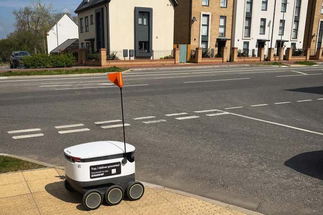 A delivery robot with an orange flag sits on a sidewalk near a road and residential buildings.