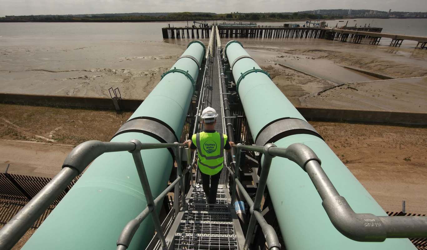A person walks along a walkway between large green pipes at a water treatment facility near a river.