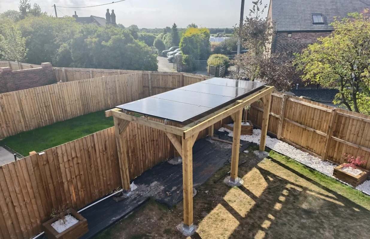 A solar panel structure on top of wooden gazebo in a fenced backyard with green grass and gravel pathways.