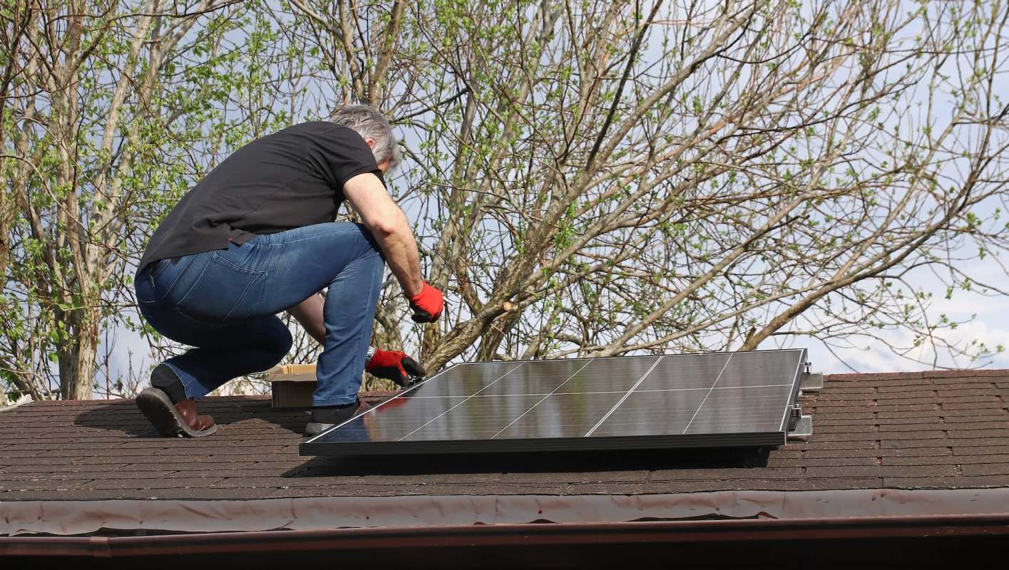 A person adjusts a solar panel on a roof with trees in the background.