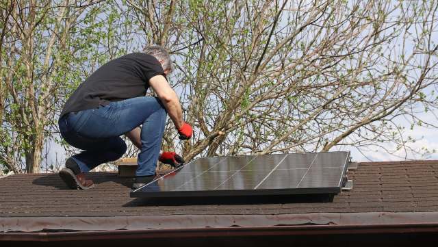 A person adjusts a solar panel on a roof with trees in the background.