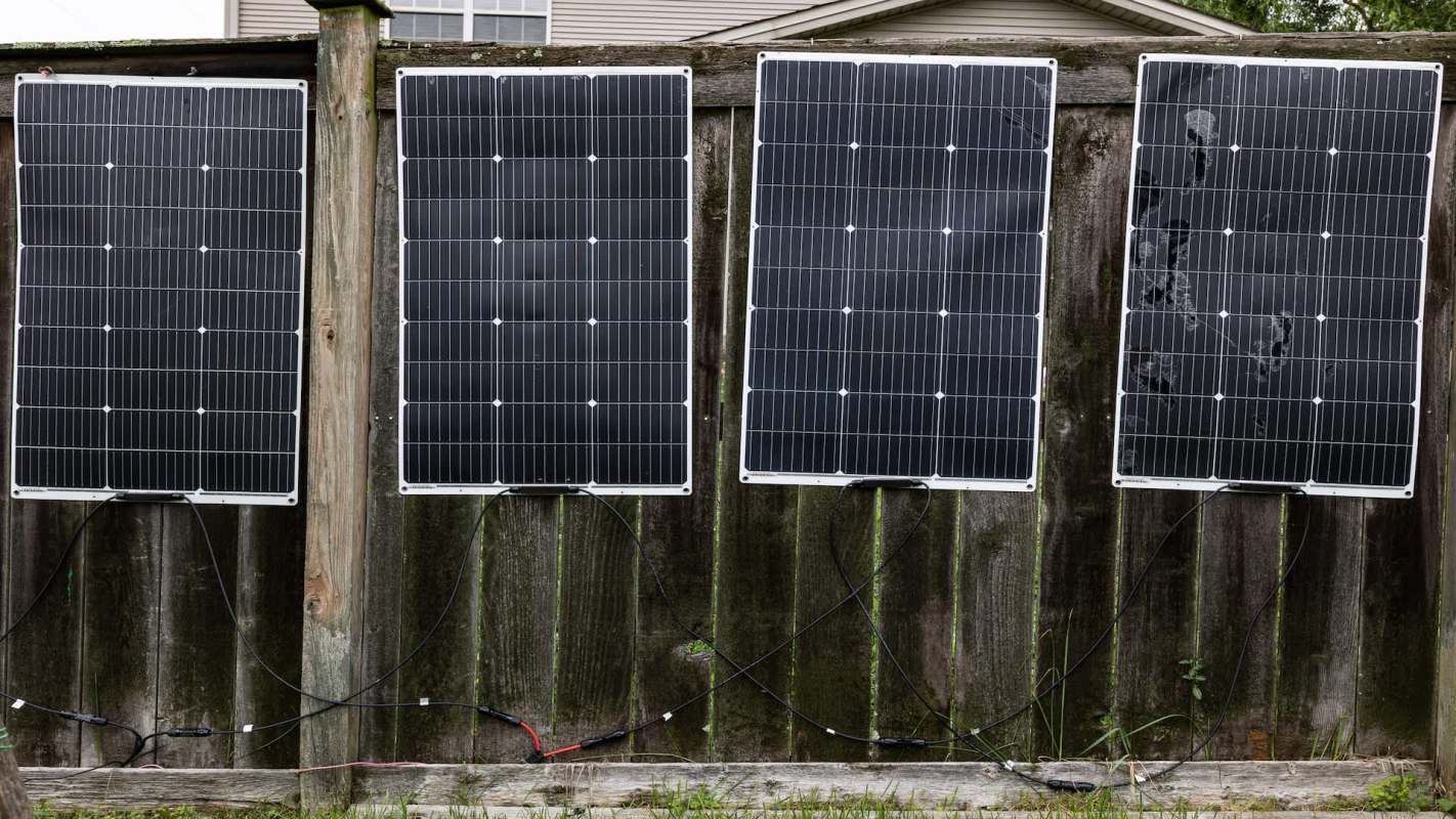 Four solar panels mounted on a wooden fence, connected by electrical wires.