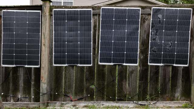 Four solar panels mounted on a wooden fence, connected by electrical wires.