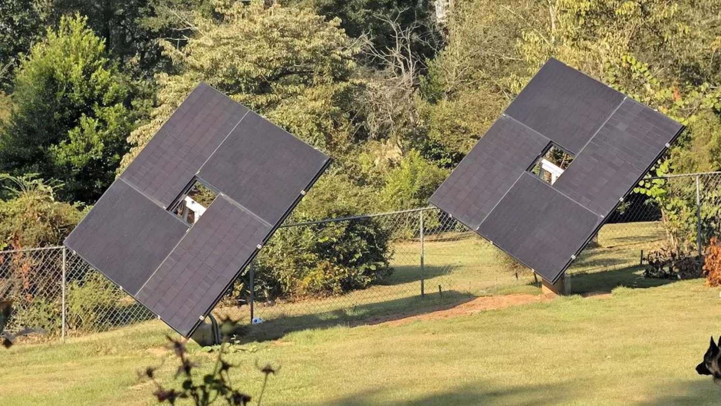 Two solar panels tilted in a grassy area with trees and a fence in the background.