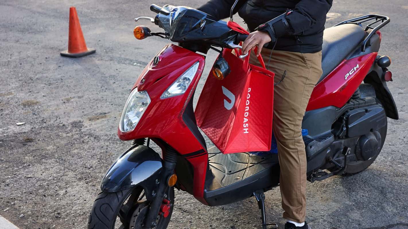 A person holds a red DoorDash delivery bag while seated on a red scooter.