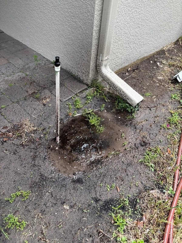 A puddle of water collects near a drainpipe on a patio with patches of grass and scattered debris.