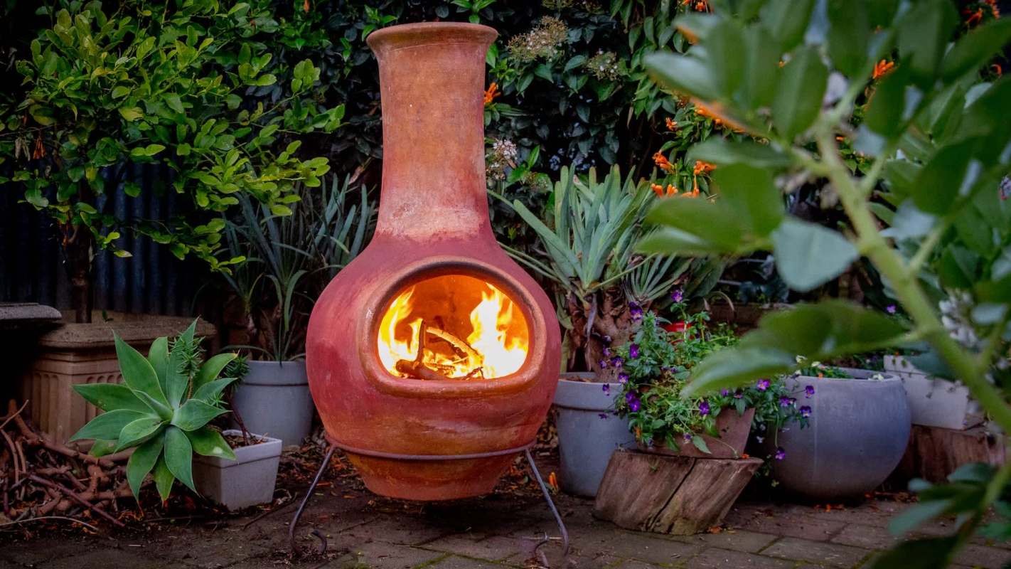 A terracotta chiminea with a fire inside, surrounded by various potted plants in a garden setting.