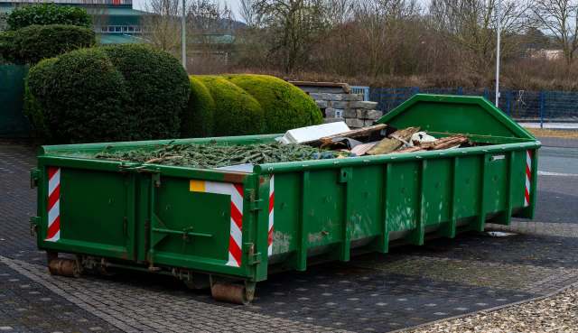 A green dumpster filled with construction debris and foliage placed on a paved area with surrounding bushes.
