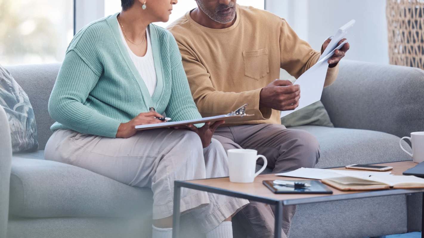 A older man and an older woman sit on a sofa, reviewing documents and taking notes with coffee mugs on the table.
