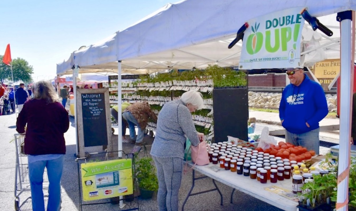 A busy farmers' market with vendors displaying jars of preserves, vegetables, and plants under tents.