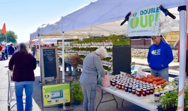 A busy farmers' market with vendors displaying jars of preserves, vegetables, and plants under tents.
