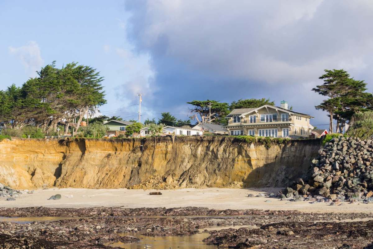 A coastal scene featuring a cliff with houses and trees above a sandy beach and rocky shoreline.
