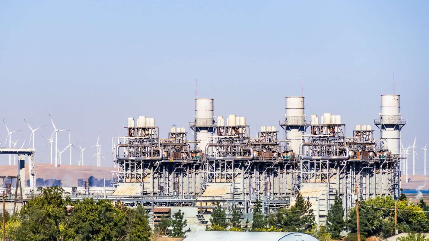 A power plant with multiple turbine structures and wind turbines in the background.