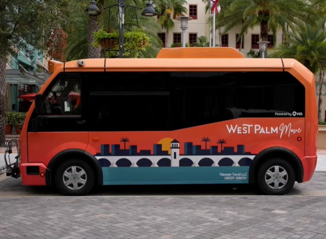 An orange bus with "West Palm Move" branding parked on a paved area surrounded by palm trees.