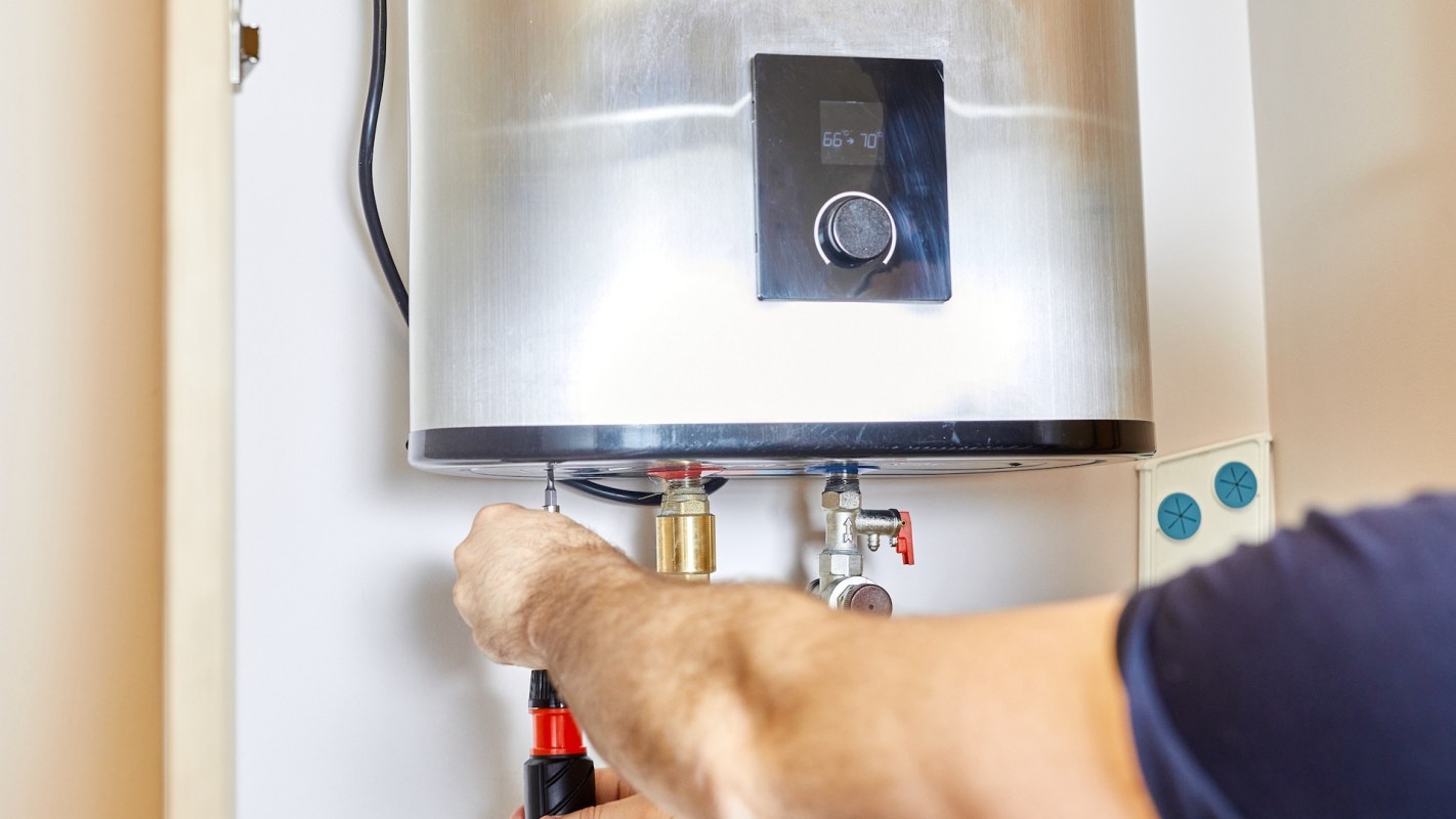 A person uses a screwdriver to work on a water heater mounted on a wall.