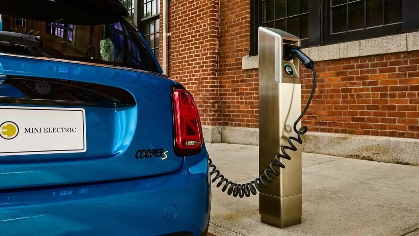 A blue electric vehicle is parked next to a curbside charging station against a brick wall.