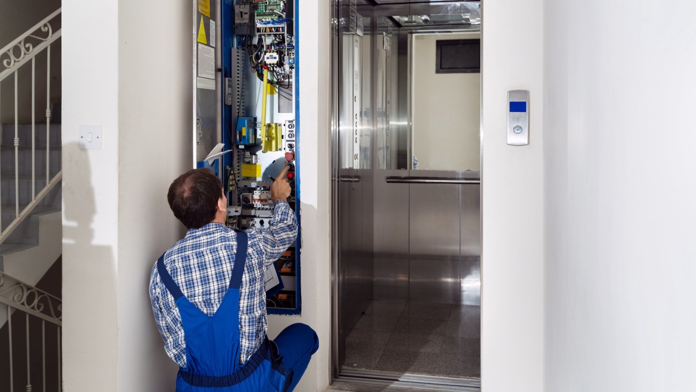 A technician works on an elevator control panel next to an open elevator door.