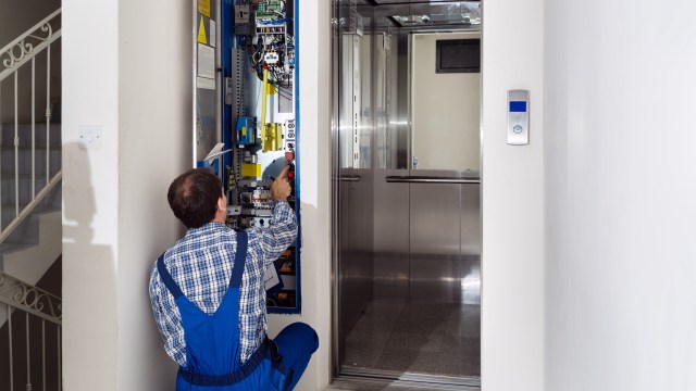 A technician works on an elevator control panel next to an open elevator door.