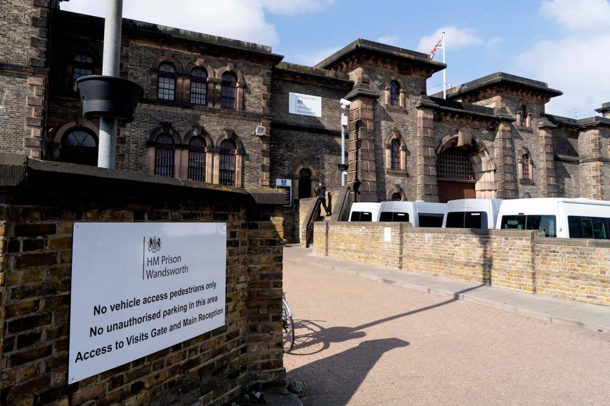 A brick prison building with a sign indicating no vehicle access and several white vans parked nearby.